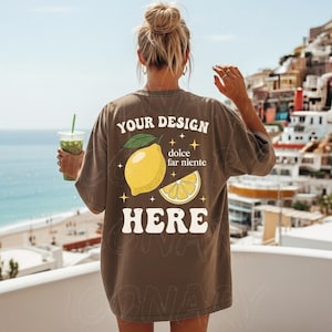 以下が含まれることがあります： レモンのグラフィックと「YOUR DESIGN HERE dolce far niente」の文字が入った茶色のTシャツ。デザインには、丸ごとのレモン、レモンスライス、星が含まれています。その人は緑色の飲み物を持っています。