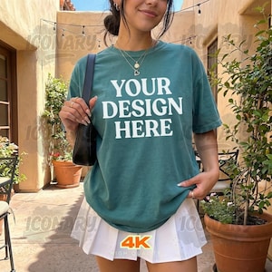 May include: A teal t-shirt with the text "YOUR DESIGN HERE" in white block letters. The shirt is paired with a white pleated skirt. The image also shows a black shoulder bag and a potted plant.