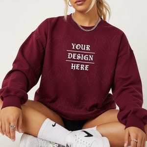 May include: A maroon sweatshirt with the text "YOUR DESIGN HERE" in white. The sweatshirt has a crew neck and long sleeves. The image shows a close-up of the sweatshirt.