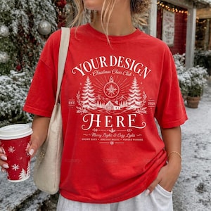 May include: A red short-sleeve t-shirt featuring a white graphic print with the text "YOUR DESIGN HERE", "Christmas Cheer Club", "EST. 2026", "Merry Nights & Cozy Lights", "SNOWY DAYS", "HOLIDAY MAGIC", and "WINTER WONDER" alongside pine trees and a cabin.