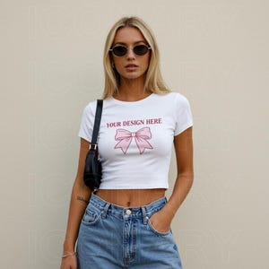 May include: White crop top with short sleeves and a pink bow design. The text "YOUR DESIGN HERE" is printed on the front of the shirt.