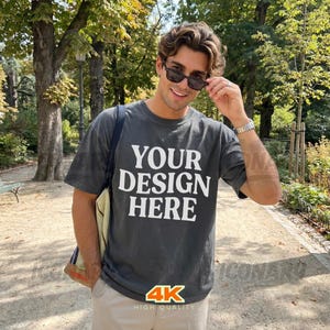 Könnte beinhalten: Dunkelgraues T-Shirt mit dem weißen Text "YOUR DESIGN HERE". Die Person trägt eine Sonnenbrille und eine beige Hose. Das Bild enthält auch ein 4K-Logo in hoher Qualität.