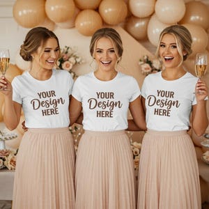 Three young women, styled as bridesmaids, wear identical white crew-neck short-sleeve t-shirts with the text "YOUR Design HERE" printed on them in a dark brown script typeface.  The t-shirts are the focus, showcasing a mockup for print design placement.  Each woman's hair is styled in a loose, elegant bun. The setting appears to be an indoor party or photoshoot, with a backdrop of champagne colored balloons and floral accents.