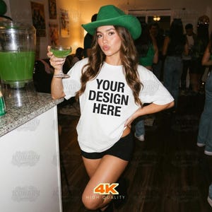 May include: A woman wearing a white t-shirt with the text "YOUR DESIGN HERE" in black, black shorts, and a green cowboy hat. She is holding a green cocktail. The image is labeled "4K HIGH QUALITY".