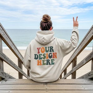 以下が含まれることがあります： レトロな文字で「YOUR DESIGN HERE」と書かれたベージュ色のパーカー。木製の階段に座っている人が着ており、背景にはビーチと海が広がっています。その人はピースサインをしています。