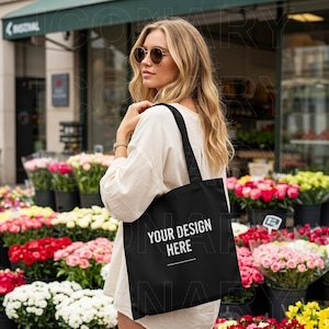 以下が含まれることがあります： 白いシャツを着てサングラスをかけた女性が、「Your Design Here」と印刷された黒いトートバッグを持っています。バッグは花屋の前にあります。