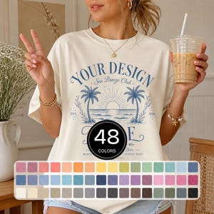 Op de afbeelding: Een crèmekleurig t-shirt met een blauwe afbeelding van palmbomen, een zon en de tekst "YOUR DESIGN Sea Breeze Club EST. 2026". Een zwarte cirkel toont de tekst "48 COLORS" boven een raster van 48 kleine gekleurde vierkantjes.