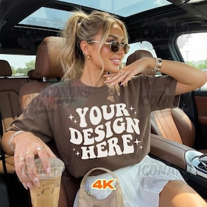 Könnte beinhalten: Ein braunes T-Shirt mit dem Text "YOUR DESIGN HERE" in Weiß, mit Sternakzenten. Das Shirt wird von einer Person in einem Auto getragen, mit Sonnenbrille und Schmuck. Eine transparente Plastikbecher mit einem Getränk wird in der Hand gehalten.
