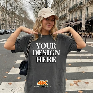 May include: Dark gray t-shirt with the text "YOUR DESIGN HERE" in large white letters. The shirt also has a "4K HIGH QUALITY" logo. The person in the image is wearing a beige baseball cap and carrying a black purse.