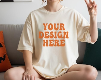 May include: Cream-colored t-shirt with the text "YOUR DESIGN HERE" in orange. The shirt has short sleeves and a relaxed fit. The image shows a person wearing the shirt while sitting on a gray couch.
