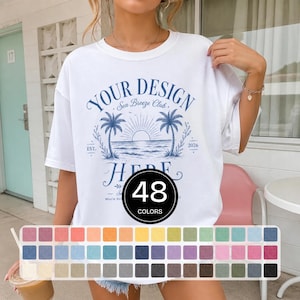 May include: A white short-sleeve t-shirt featuring a blue graphic print of palm trees, a sun, and the ocean. The text reads "YOUR DESIGN HERE", "Sea Breeze Club", "EST. 2026". A circular overlay displays "48 COLORS" above a grid of 48 different coloured squares.