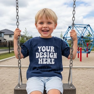 May include: A young child wearing a navy blue t-shirt with the text "YOUR DESIGN HERE" in white, sitting on a swing set. The child is smiling, with a playground and houses in the background.
