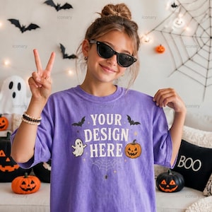 May include: A purple short-sleeve t-shirt featuring the white text 'YOUR DESIGN HERE' surrounded by black and orange Halloween graphics including a ghost, a jack-o'-lantern, bats, stars, and a spiderweb design printed on the front center.