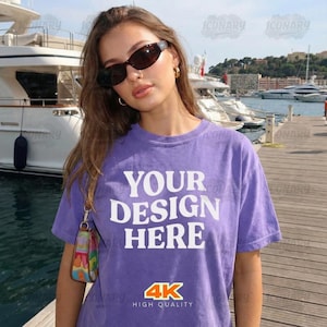 Peut inclure: Un t-shirt violet avec le texte "YOUR DESIGN HERE" en lettres blanches. Le t-shirt comporte également le texte "4K HIGH QUALITY" en orange et blanc. La personne porte des lunettes de soleil et un petit sac à main coloré.