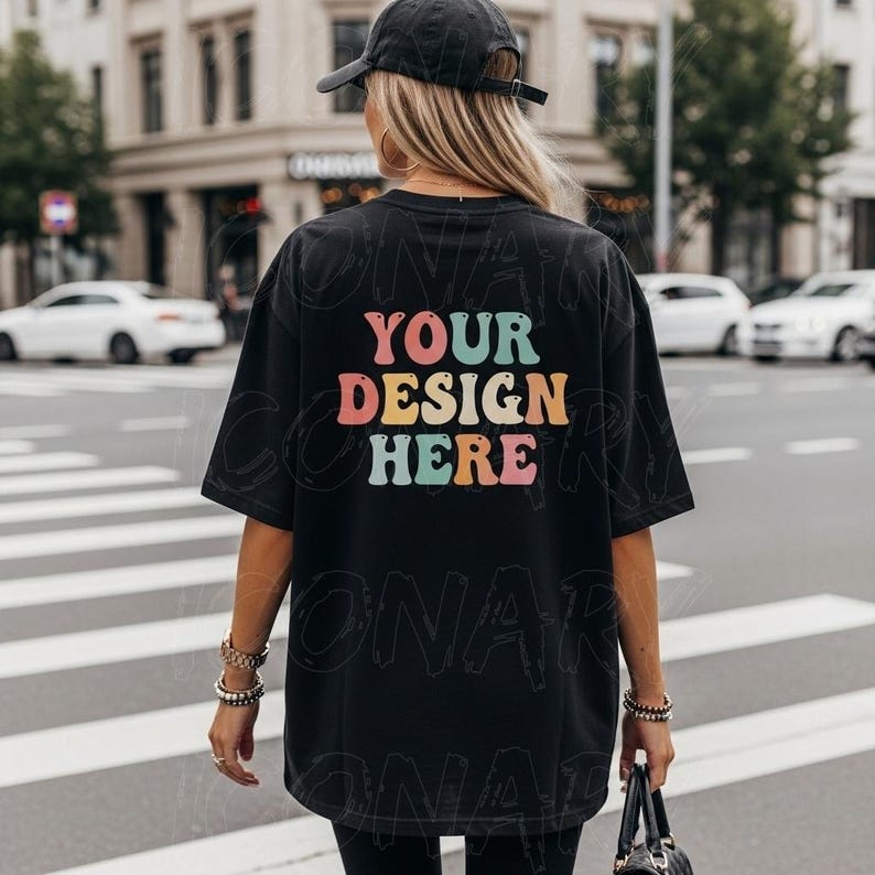May include: Black oversized t-shirt with the text "YOUR DESIGN HERE" in colorful retro font. The model is wearing a black cap and holding a black purse. The background shows a city street with crosswalk.