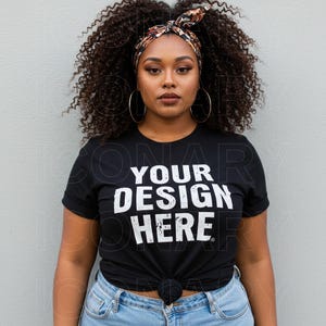 A plus-size Black fashion model with curly, voluminous dark brown hair styled with a patterned headband, is pictured against a light gray wall.  She is wearing a white t-shirt that displays the words "YOUR DESIGN HERE" in a bold, black sans-serif font with textural variations, as if distressed or painted on.  She wears large hoop earrings and light wash blue jeans. Her makeup is natural, with a warm brown lipstick and subtle eye definition.