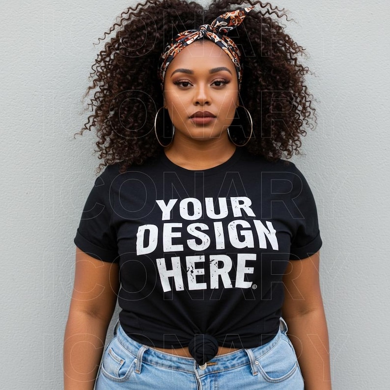 A plus-size Black fashion model with curly, voluminous dark brown hair styled with a patterned headband, is pictured against a light gray wall.  She is wearing a white t-shirt that displays the words "YOUR DESIGN HERE" in a bold, black sans-serif font with textural variations, as if distressed or painted on.  She wears large hoop earrings and light wash blue jeans. Her makeup is natural, with a warm brown lipstick and subtle eye definition.