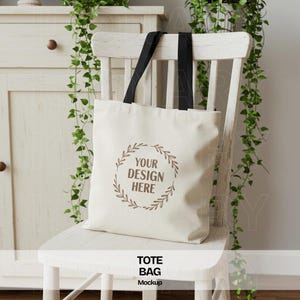 May include: A white canvas tote bag with black handles. The bag has a brown wreath design with the text "YOUR DESIGN HERE" in the center. The bag is sitting on a white chair.