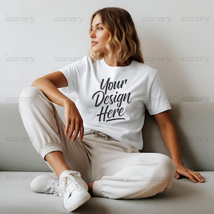 Op de afbeelding: Een vrouw draagt een wit T-shirt met de tekst "Your Design Here" in zwarte schrift. Ze zit op een grijze bank met haar benen gekruist en draagt witte sneakers.