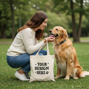 May include: A woman in a white sweater and blue jeans is crouching down next to a golden retriever dog. She is holding a white tote bag with the text "YOUR DESIGN HERE" printed on it.