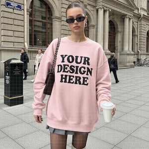 Puede incluir: Sudadera rosa claro con el texto "YOUR DESIGN HERE" en negro. La sudadera de manga larga tiene cuello redondo. La persona lleva gafas de sol y sostiene una taza de café.