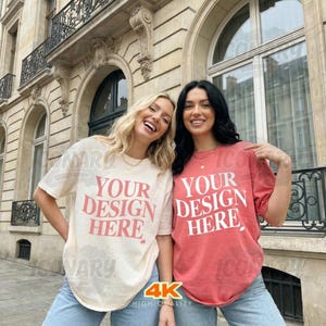 May include: Two women wearing light-colored t-shirts with the text "YOUR DESIGN HERE" in a red font. The shirts are paired with blue jeans. The background features a stone building with arched windows and decorative ironwork.