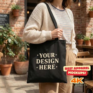 May include: A black tote bag with the text "YOUR DESIGN HERE" in white. The bag is being held by a person. The bag has a black strap and a white strap. The image also includes the text "BEST APPAREL MOCKUPS!" and "4K HIGH QUALITY".