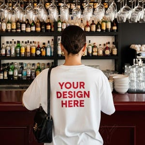 Puede incluir: Una camiseta blanca con el texto "YOUR DESIGN HERE" en rojo. La persona está de pie en un bar con botellas de licor y vasos colgando encima. Un bolso negro está en el hombro de la persona.
