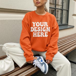 Könnte beinhalten: Ein orangefarbenes Sweatshirt mit dem weißen Text "YOUR DESIGN HERE", kombiniert mit cremefarbenen Hosen und weißen Turnschuhen mit blauen Akzenten. Eine weiße Handtasche liegt auf einer Holzbank.
