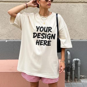 Puede incluir: Camiseta beige de gran tamaño con el texto negro "YOUR DESIGN HERE". La modelo lleva un pantalón corto a cuadros rojos y blancos y un bolso negro. La persona lleva joyas doradas y gafas de sol.
