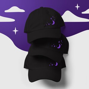 Könnte beinhalten: Drei schwarze Baseballkappen, die übereinander gestapelt sind. Jede Kappe hat eine gestickte, violette Mondsichel und Sterne auf der Vorderseite.
