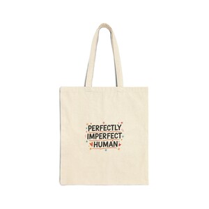 May include: A cream-colored tote bag with a simple design. The bag features the text "PERFECTLY IMPERFECT HUMAN" in a colorful, playful font. The bag has two long handles for carrying.