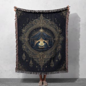 May include: A dark blue woven throw blanket with a gold ornate design. The center features a circular frame with a depiction of two figures in a tent. The blanket has a fringed edge and is held up by a person.