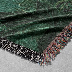 May include: A woven throw blanket with a dark green botanical pattern. The blanket has a black and white fringe on one side and a multicolored fringe on the other. The design features large leaves and a marbled effect.