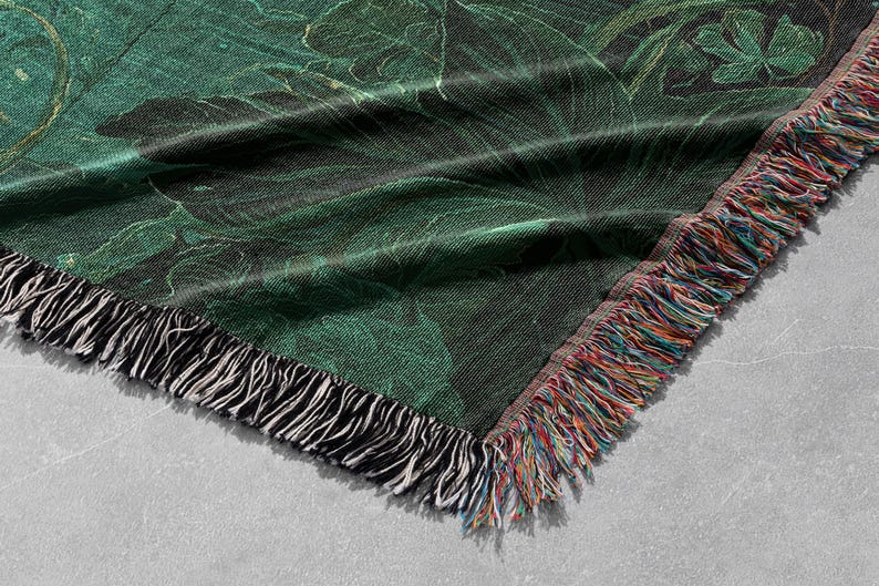 May include: A woven throw blanket with a dark green botanical pattern. The blanket has a black and white fringe on one side and a multicolored fringe on the other. The design features large leaves and a marbled effect.