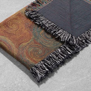 May include: A woven throw blanket with a swirling pattern in shades of orange, yellow, and green. The blanket has a dark gray backing and black and white fringe trim. The blanket is folded over on a light gray surface.