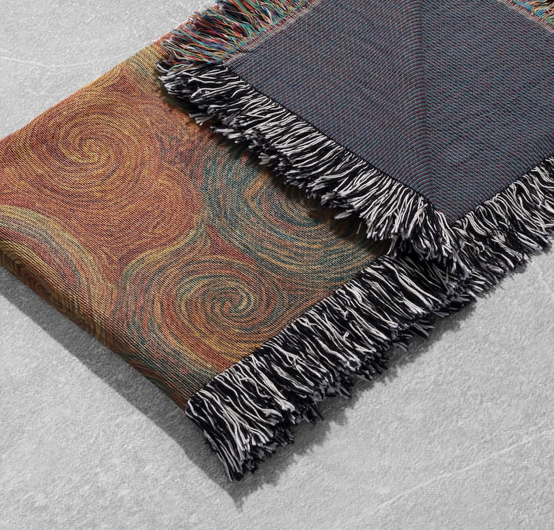 May include: A woven throw blanket with a swirling pattern in shades of orange, yellow, and green. The blanket has a dark gray backing and black and white fringe trim. The blanket is folded over on a light gray surface.