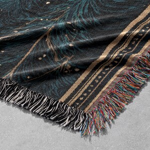 May include: A woven throw blanket with a dark teal and black pattern, accented with gold and white details. The blanket has a fringed edge with a mix of colorful threads. The design is intricate and detailed.
