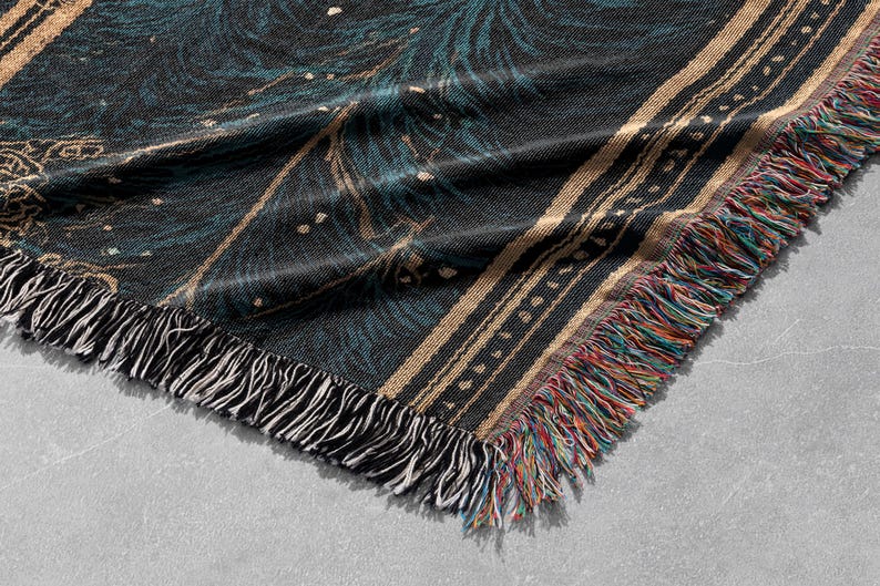 May include: A woven throw blanket with a dark teal and black pattern, accented with gold and white details. The blanket has a fringed edge with a mix of colorful threads. The design is intricate and detailed.