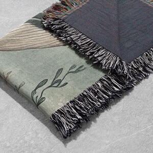 May include: A woven throw blanket with a green and brown floral pattern and a black fringe. The blanket is folded in half and the fringe is visible on both sides.