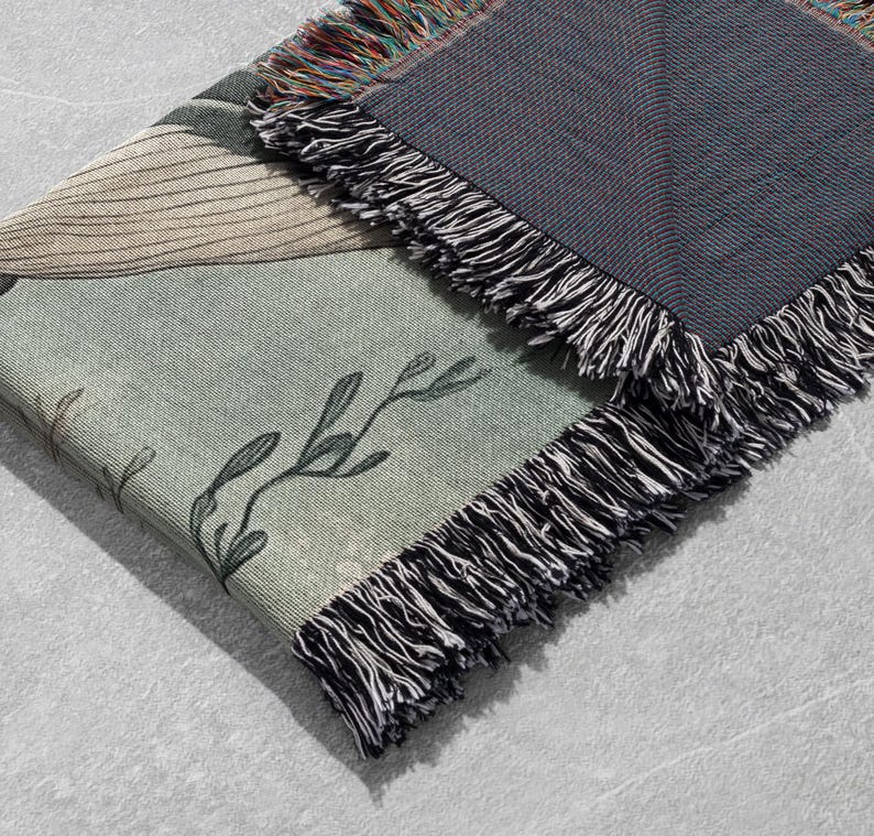 May include: A woven throw blanket with a green and brown floral pattern and a black fringe. The blanket is folded in half and the fringe is visible on both sides.