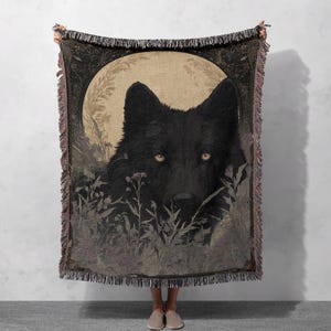 May include: A woven throw blanket depicting a black wolf with yellow eyes, set against a full moon and floral details. The blanket has a fringed edge and a detailed design. The colour scheme includes black, beige, and muted purple, creating a striking visual.