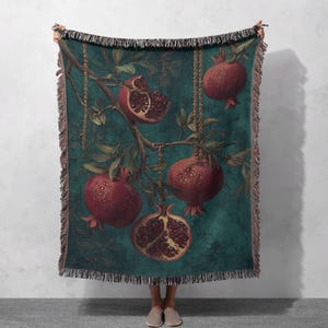 May include: A woven throw blanket featuring a botanical illustration of pomegranates on a teal background. The design includes ripe, red pomegranates hanging from branches with green leaves. The blanket has a fringed edge.