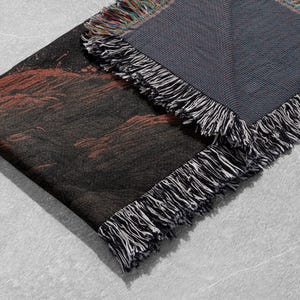 May include: A woven throw blanket with a dark color palette. The blanket features a black and red design on one side and a gray reverse side. The edges are finished with a fringe in black, white, and multicolored threads.