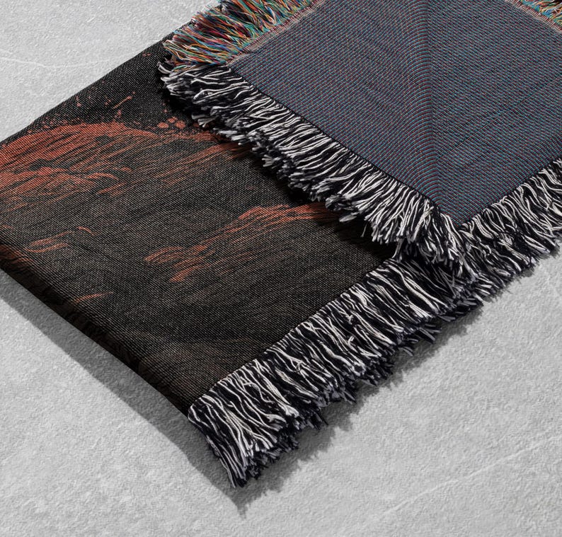 May include: A woven throw blanket with a dark color palette. The blanket features a black and red design on one side and a gray reverse side. The edges are finished with a fringe in black, white, and multicolored threads.