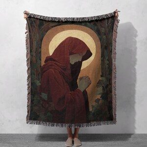 May include: Woven throw blanket featuring a religious figure in a red robe, hands clasped in prayer. The artwork is set against a golden halo and a backdrop of stylized trees and foliage. The blanket has a fringed edge.