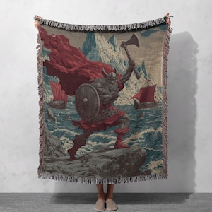Puede incluir: Manta tejida que representa a un guerrero vikingo con hacha y escudo, capa roja y casco con cuernos. El diseño incluye barcos vikingos en el mar, montañas y una paleta de colores azul y beige. La manta tiene un borde con flecos.