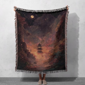 Könnte beinhalten: Gewebte Decke mit einer Landschaft mit einer Pagode, Bergen und einem Sternenhimmel. Das Design verwendet Rot-, Orange- und Brauntöne, mit einem Vollmond. Die Decke hat einen Fransenrand.
