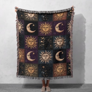 May include: A woven throw blanket featuring a repeating pattern of suns and crescent moons in squares. The blanket has a fringed edge and is primarily in shades of gold, purple, and black. The celestial design adds a mystical touch to home decor.