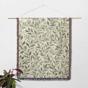 Op de afbeelding: Een rechthoekige wandhanger met een botanisch bladerpatroon in groen en beige. De bladeren zijn gerangschikt in een vloeiend, naturalistisch ontwerp. De randen hebben een gefranjerde rand. De wandhanger is opgehangen aan een houten roede.